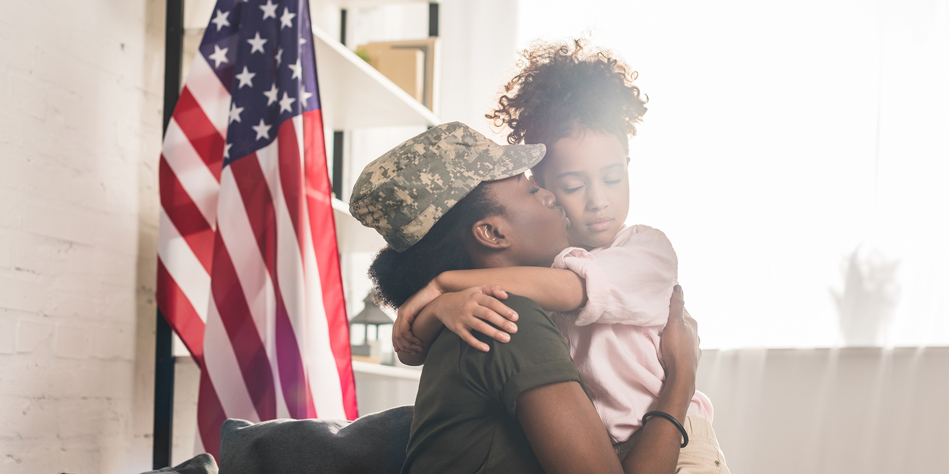 African America Woman in camouflage Army uniform hugging her daughter with American Flag in background